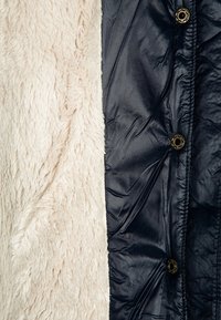 Miękka kremowa polarowa tkanina obok ciemnogranatowej błyszczącej pikowanej tkaniny z trzema metalowymi zatrzaskami.