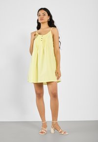 Gelbes ärmelloses Kleid mit durchgehender Knopfleiste, gerafftem Stoff und knielangem Schnitt. Modell kombiniert mit beigefarbenen, riemchen Sandalen mit Blockabsatz.