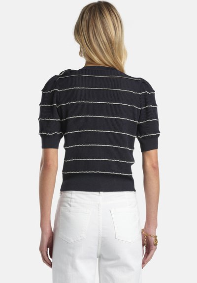 Femeie blondă purtând un tricou negru cu mâneci scurte, cu dungi orizontale albe și blugi albi cu talie înaltă, cu fața spre spate.