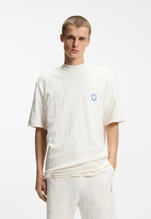 Jonge man met kort haar, gekleed in een wit textuur kortermig shirt en bijpassende broek met een klein blauw logo op de borst en dij.