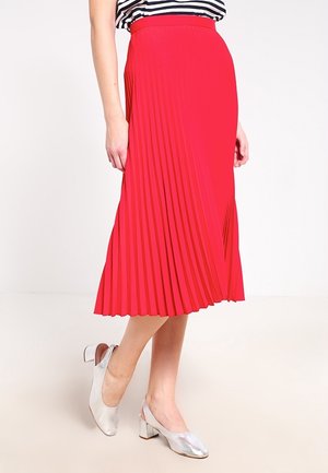 Mujer con falda midi roja plisada, blusa de rayas y zapatos slingback plateados de tacón bajo, de pie frente a un fondo sencillo.