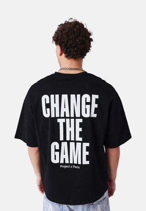 Uomo con capelli ricci che indossa una maglietta nera con la scritta bianca in grassetto "CHANGE THE GAME" e un testo più piccolo "Project x Paris" sul retro.