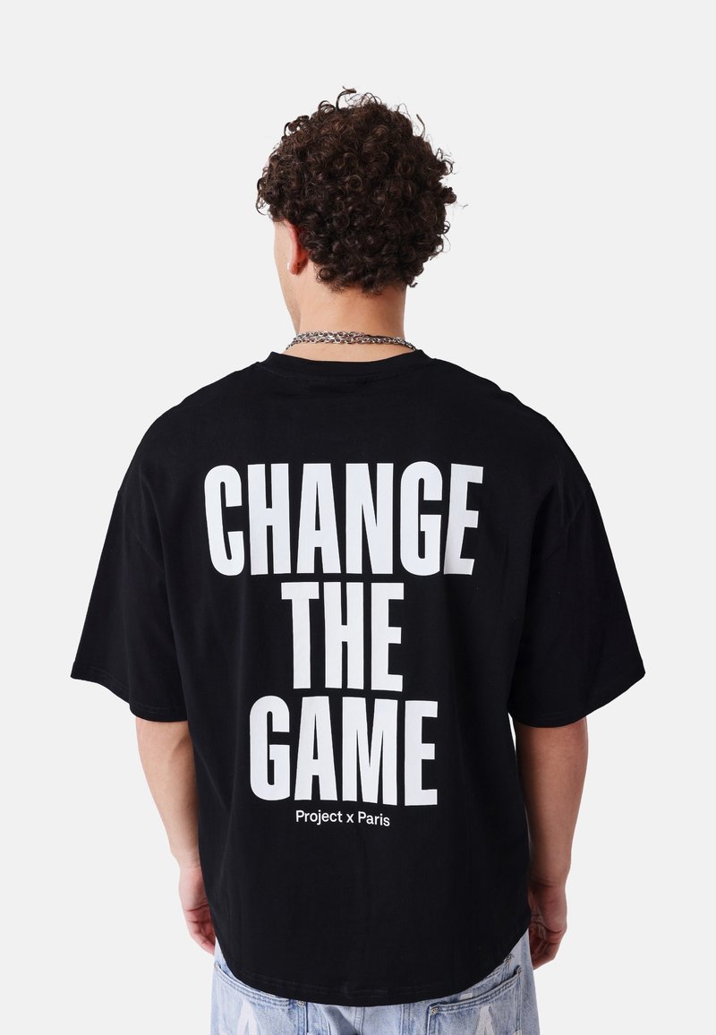 Homme aux cheveux bouclés portant un t-shirt noir avec un texte blanc en gras « CHANGE THE GAME » et un texte plus petit « Project x Paris » au dos.