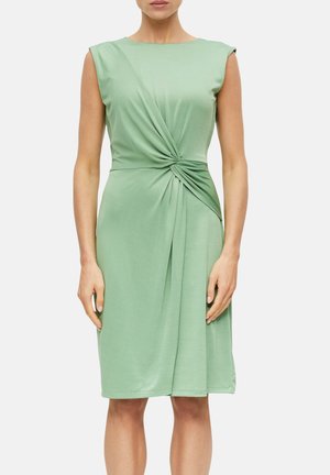 Femme portant une robe sans manches vert clair avec un détail froncé torsadé à la taille, debout devant un fond blanc uni.
