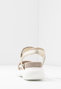 Zadní pohled na sandál s zlatým kovovým páskem, béžovými semišovými akcenty a bílou texturovanou podrážkou. Design zahrnuje nastavitelné popruhy.