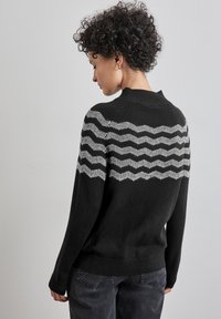 Schwarzer Pullover mit einem wellenförmigen weißen Muster über dem oberen Rücken und den Schultern. Er hat einen hohen Kragen sowie Rippbündchen an den Ärmeln und am Saum.
