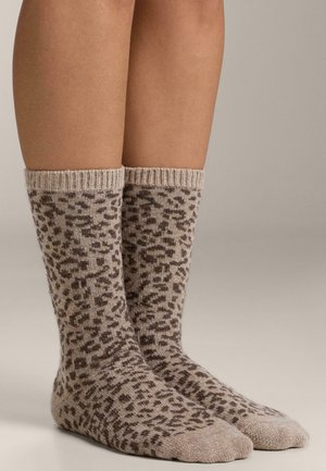 Noge u bež čarapama srednje dužine s tamno smeđim leopardovim uzorkom, stoje na neutralnoj pozadini.