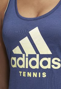 Mörkblå linne i andningsbart material, med en stor gul adidas-logotyp och texten "TENNIS" i fetstil, i kontrasterande färger.