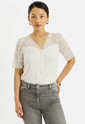 Mujer con blusa blanca de encaje de manga corta con bordes ondulados y botones, combinada con vaqueros grises descoloridos de tiro alto.