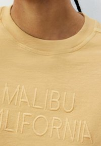 Žlutá mikina s texturovaným, vyšívaným designem "MALIBU CALIFORNIA" na přední straně. Má žebrovaný límec a měkkou tkaninu.