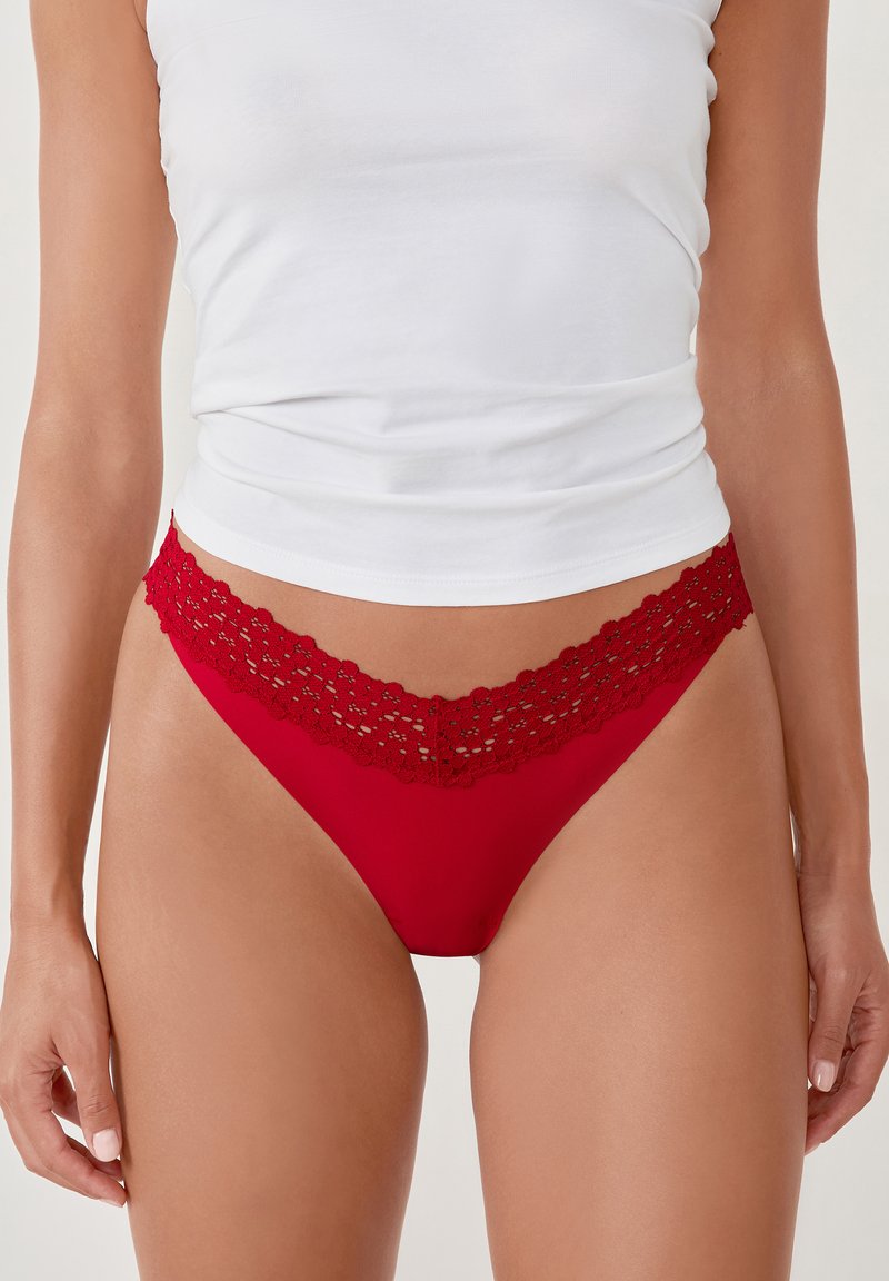 Femme portant des sous-vêtements de style bikini en dentelle rouge et un débardeur blanc, montrée de mi-cuisse à juste en dessous de la poitrine.