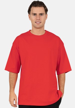 Joven con cabello castaño que lleva una camiseta roja de manga corta y holgada, de pie frente a un fondo claro y liso.