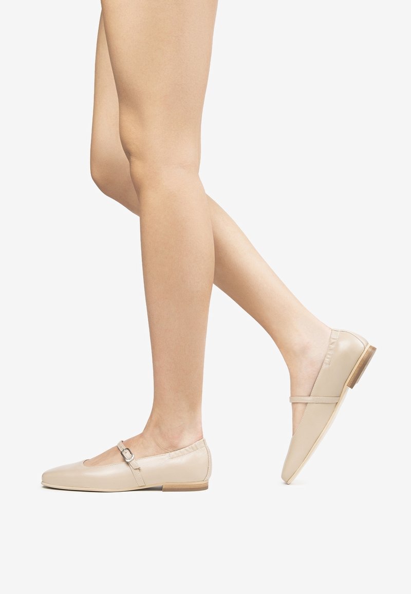 Jambes nues portant des chaussures plates Mary Jane en cuir beige avec une petite boucle et un petit talon en bois, un pied légèrement levé derrière l'autre.