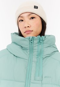 Chaqueta acolchada en color azul verdoso con cuello alto, cremallera de dos vías y costuras sutiles. Gorro de canalé color crema con una etiqueta, usado por una persona.