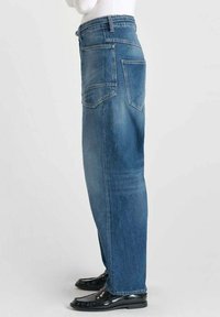 Jean droit en denim bleu clair avec une taille haute, cinq poches et des zones légèrement décolorées. Porté avec des chaussures noires, montrant le profil latéral.