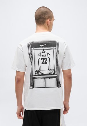Mand iført hvid T-shirt med sort-hvid grafik af en basketballtrøje, sko og bold i et omklædningsrum.