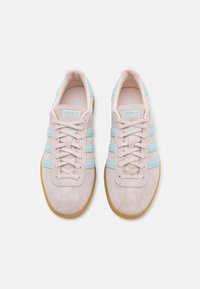 Ett par ljusrosa mockaskor från Adidas med pastellblå ränder och gummiklädda sulor, sedd ovanifrån mot en vit bakgrund.