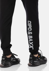 Schwarze Jogginghosen mit weißem "CIPO & BAxx"-Text an der Seite, mit schlanker Passform und elastischen Bündchen. Kombiniert mit schwarzen Sneakers mit "AIR"-Markierung.