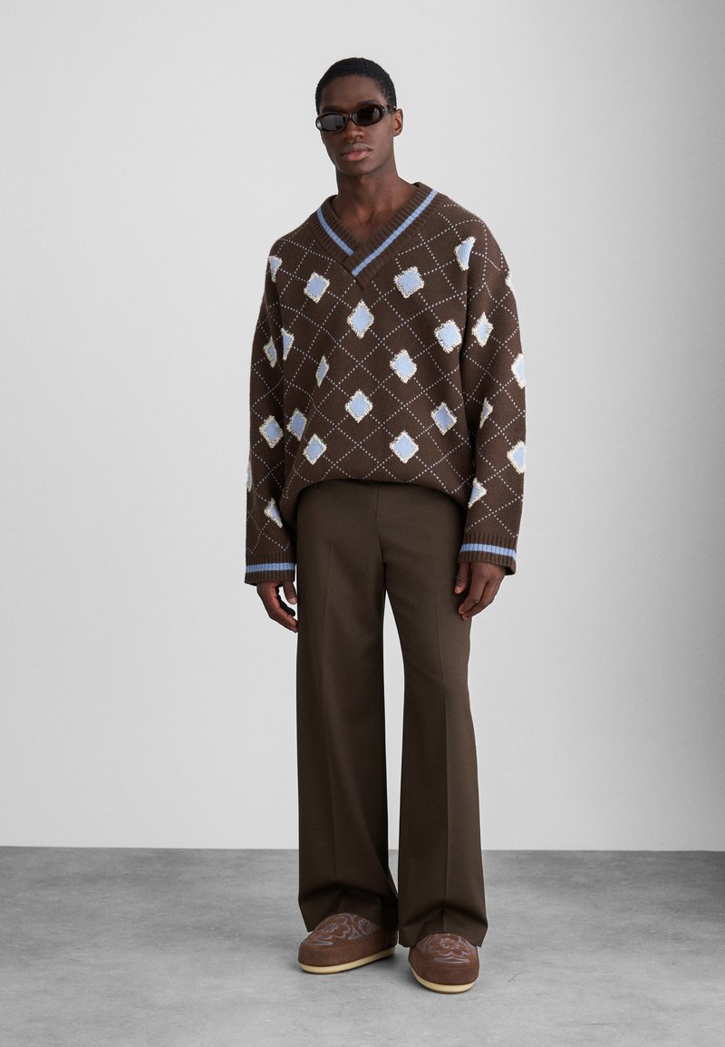 Bruine gebreide trui met geometrische patronen, V-hals en blauwe accenten, gecombineerd met bruine wijde broek en bruine slippers met patroon.