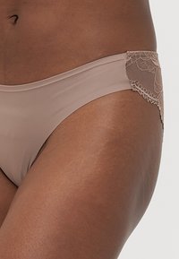 Nude-colored bikini bottom with smooth fabric and lace detailing on the waist, featuring a floral pattern and scalloped edges.