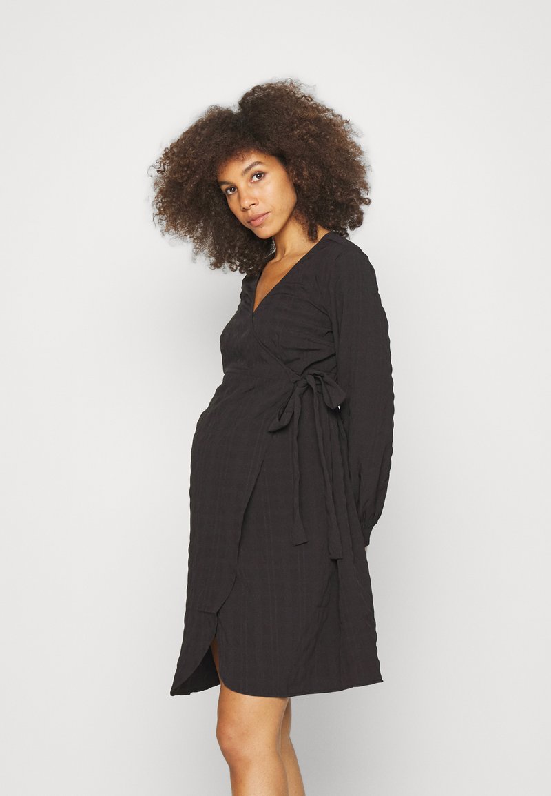 Vestido preto envolvente com mangas longas, tecido texturizado, decote em V e um laço na cintura. Apresenta uma fenda lateral para um detalhe adicional.