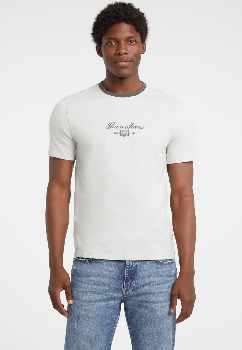 Camiseta de color gris claro con un cuello en contraste de color gris oscuro, que presenta el logo "Guess Jeans" en negro, mangas cortas, diseño ajustado y estilo casual.