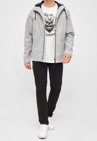 Hellgraue Softshell-Jacke mit Kapuze, schwarzen Akzenten und einem Reißverschluss vorne, getragen über einem weißen T-Shirt mit Grafik, kombiniert mit schwarzen Hosen und weißen Turnschuhen.