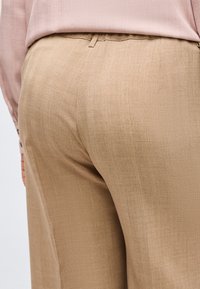 Μπεζ ραμμένα παντελόνια, φορεμένα με ροζ πουκάμισο με μακριά μανίκια, μπαλανρισμένο μέσα, που δείχνει την υφή του υφάσματος και την εφαρμογή από πίσω.