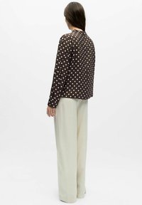 Kvinne med langt hår iført en brun, langermet topp med polkaprikkmønster og løse, hvite bukser, som står med ryggen mot på en hvit bakgrunn.