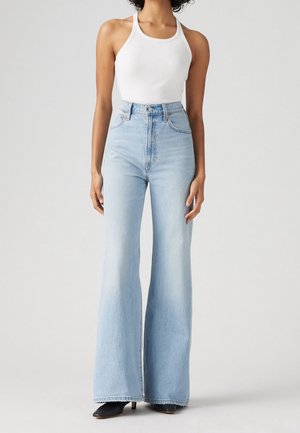 Femme portant un jean évasé taille haute bleu clair, un haut blanc sans manches en maille côtelée et des chaussures à talons noires sur fond uni.