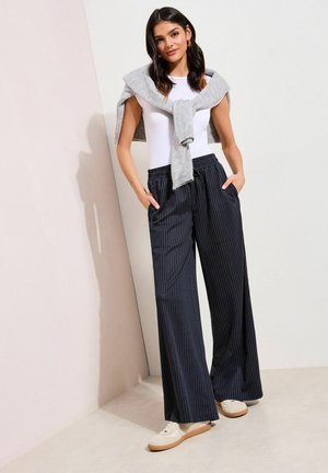 Žena v bielom tielku bez rukávov, sivom svetri prehodenom cez ramená, námornícky modrých širokých nohaviciach s tenkými pásikmi a bielych teniskách, stojaca v interiéri.