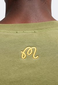 Vue de dos d'une personne portant une chemise vert olive avec un petit motif jaune brodé près du col au niveau du cou.