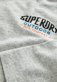 Grijze katoenen T-shirt met opvallende zwarte, blauwe en oranje tekst "SUPERDRY OUTDOOR MFG. COMPANY" over de borst. Zachte, gestructureerde stof.