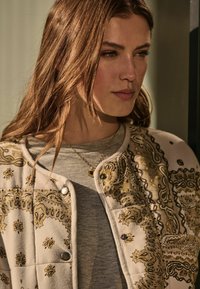 Beige gewatteerde jas met een ingewikkeld gouden paisleypatroon, zilveren drukknoopjes, gedragen over een grijs shirt, met een subtiele ronde gouden pendant ketting.