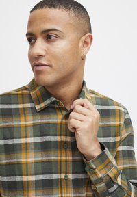 Skjorte i plaid mønster i grøn, orange og beige; lavet af tekstureret stof med knapper og en klassisk krave; viser en hånd der griber fat i kraven.