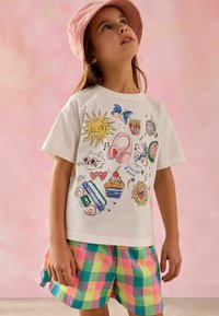 Biała koszulka z kolorowymi ilustracjami, w tym słońcem, słuchawkami i jedzeniem, w połączeniu z kratkowymi pastelowymi szortami. Różowa kapelusz typu bucket.