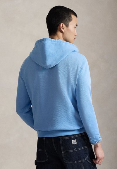 Muškarac obučen u svijetloplavu hoodicu i tamne traperice s džepovima na stražnjoj strani, stoji okrenut u stranu, na neutralnoj bež pozadini.