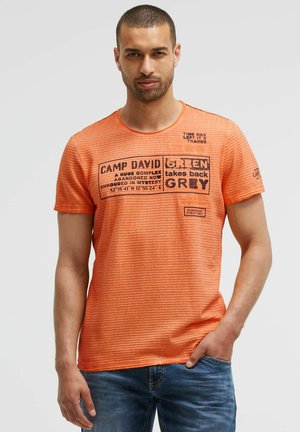 Man wearing an orange striped t-shirt with black printed text and blue jeans, standing against a plain light background.