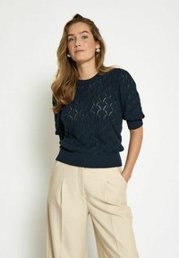 Navy knit sweater with a textured leaf pattern, short sleeves, ribbed hem, paired with beige pleated trousers.