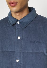 Marineblå kordfløjl skjorte med knapper og krave, udstyret med sølvfarvede trykknapper og broderet logo på brystet. Teksturdetaljer i stoffet er synlige.