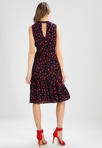 Robe sans manches en tissu bleu marine foncé avec un motif de cœurs rouges, ceinture à la taille et ourlet volanté évasé. Détail de dos en forme de clé et escarpins rouges.