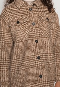 Chaqueta de camisa de cuadro marrón hecha de tela acariciada; cuenta con botones negros, dos bolsillos en el pecho y un cuello. La textura es suave y cálida.