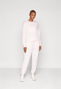 Haut à capuche rose clair avec une poche avant et un cordon de serrage, associé à un pantalon de jogging fuselé assorti. Le matériau semble doux et confortable.