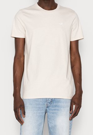 Hombre vistiendo una camiseta de manga corta beige lisa con un pequeño logo blanco en el pecho, combinada con unos jeans de color azul claro.