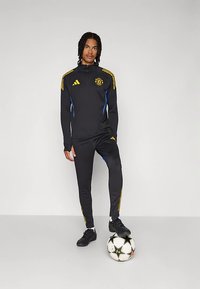 Melns sporta tērps ar rāvējslēdzēju jaku, kurai ir dzelteni un zili svītraini akcenti, un atbilstošas, šaurākas bikšu daļas. Melni sporta apavi un futbolbumba.