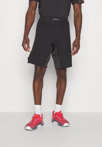 Schwarze Sportshorts mit glatter Textur, ausgestattet mit einem breiten Bund und seitlichen Einsätzen. Kombiniert mit lebhaft roten und grauen Sneakers.