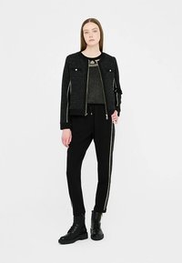 Black bomber jacket with gold hardware, paired with a dark top and black trousers featuring a side stripe; model wearing black ankle boots.