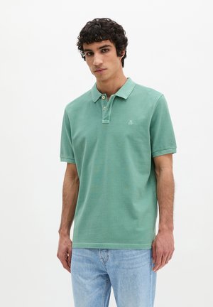 Jeune homme aux cheveux bouclés portant un polo vert à manches courtes et un jean bleu clair, sur un fond blanc uni.