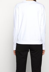 Vit sweatshirt med sänkt axel, ribbade muddar och nederkant. Bakvy visar en slät yta utan synliga mönster eller accenter. Svarta byxor bärs.