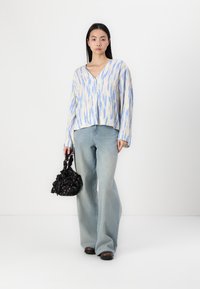 Leichte, gemusterte Bluse mit blauen und cremefarbenen Designs, kombiniert mit hellen, weit geschnittenen Jeans und einer schwarzen strukturierten Tasche mit floralen Akzenten.